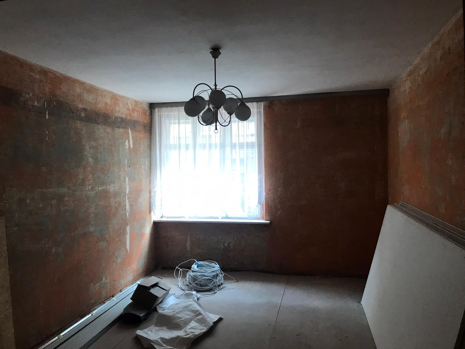 Pokój w trakcie remontu: ściany z nierówną powierzchnią, widoczne ślady po starych warstwach, przygotowane płyty gipsowo-kartonowe oparte o ścianę, zwój kabli elektrycznych i metalowe profile...