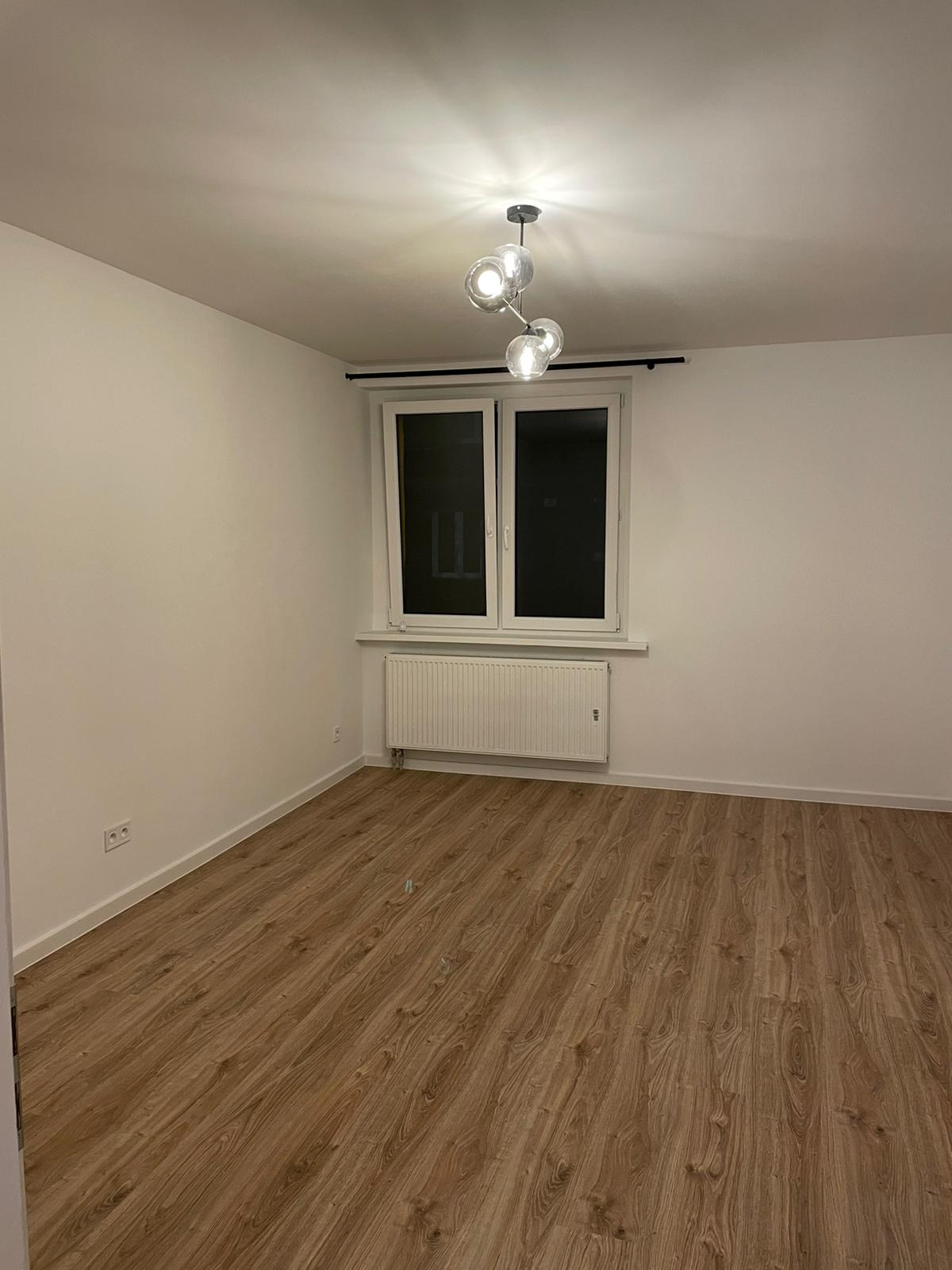 Pusty pokój z panelami podłogowymi w odcieniach brązu, białymi ścianami i sufitem, grzejnikiem pod oknem i nowoczesną lampą sufitową z kulistymi kloszami.