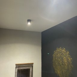 ELEWACJE I REMONTY ADAM CYRANIAK - Częściowe ujęcie wnętrza: ciemna ściana z dekoracyjnym, złotym ornamentem w stylu mandali, jasna ściana z oknem w żaluzjach, punktowe oświetlenie sufitowe.