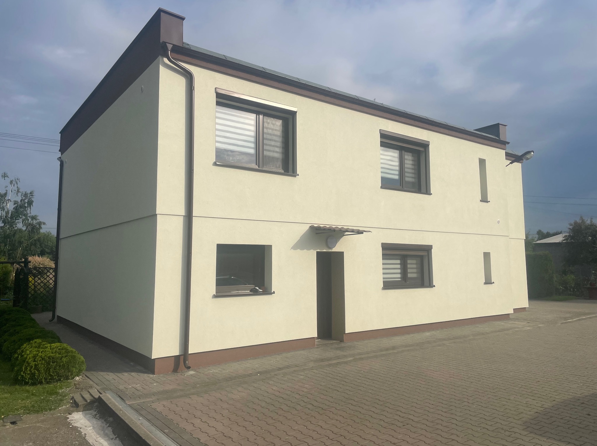 Dwukondygnacyjny dom z jasną elewacją, brązowymi obramowaniami okien i dachu, widoczny z poziomu brukowanego podjazdu w słoneczny dzień.