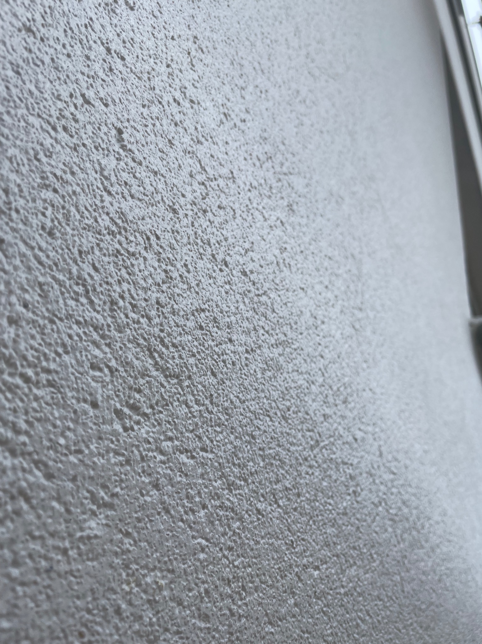 Jasnoszara, chropowata faktura ściany zewnętrznej z widocznym fragmentem ramy okiennej po prawej stronie.