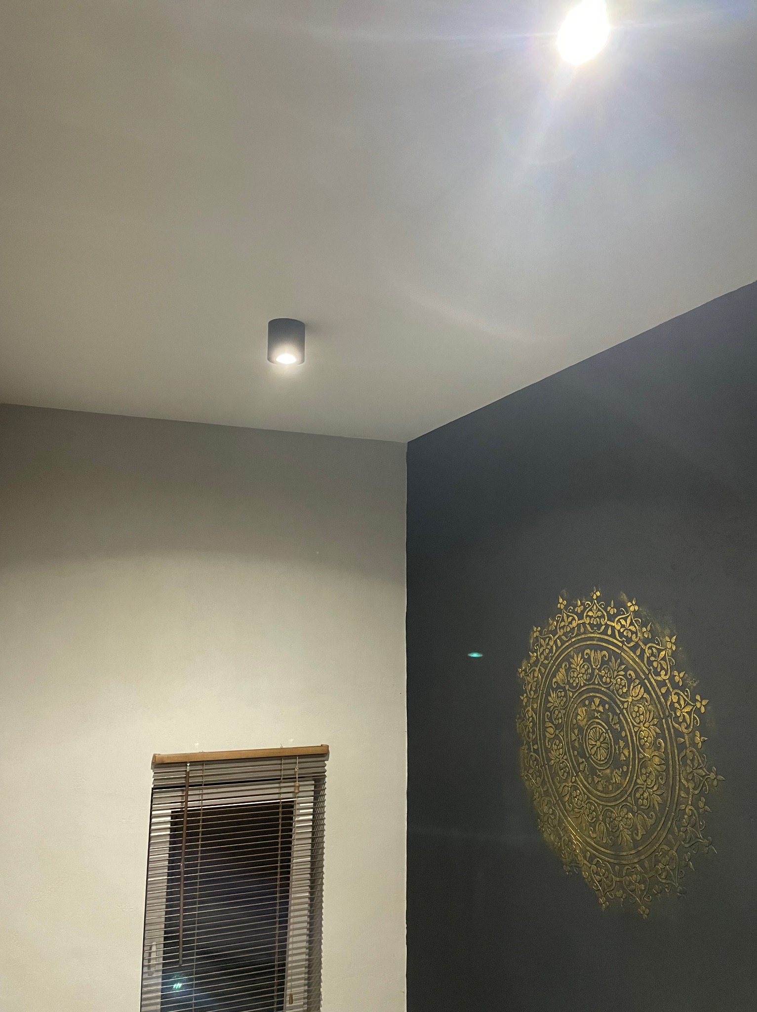 Częściowe ujęcie wnętrza: ciemna ściana z dekoracyjnym, złotym ornamentem w stylu mandali, jasna ściana z oknem w żaluzjach, punktowe oświetlenie sufitowe.