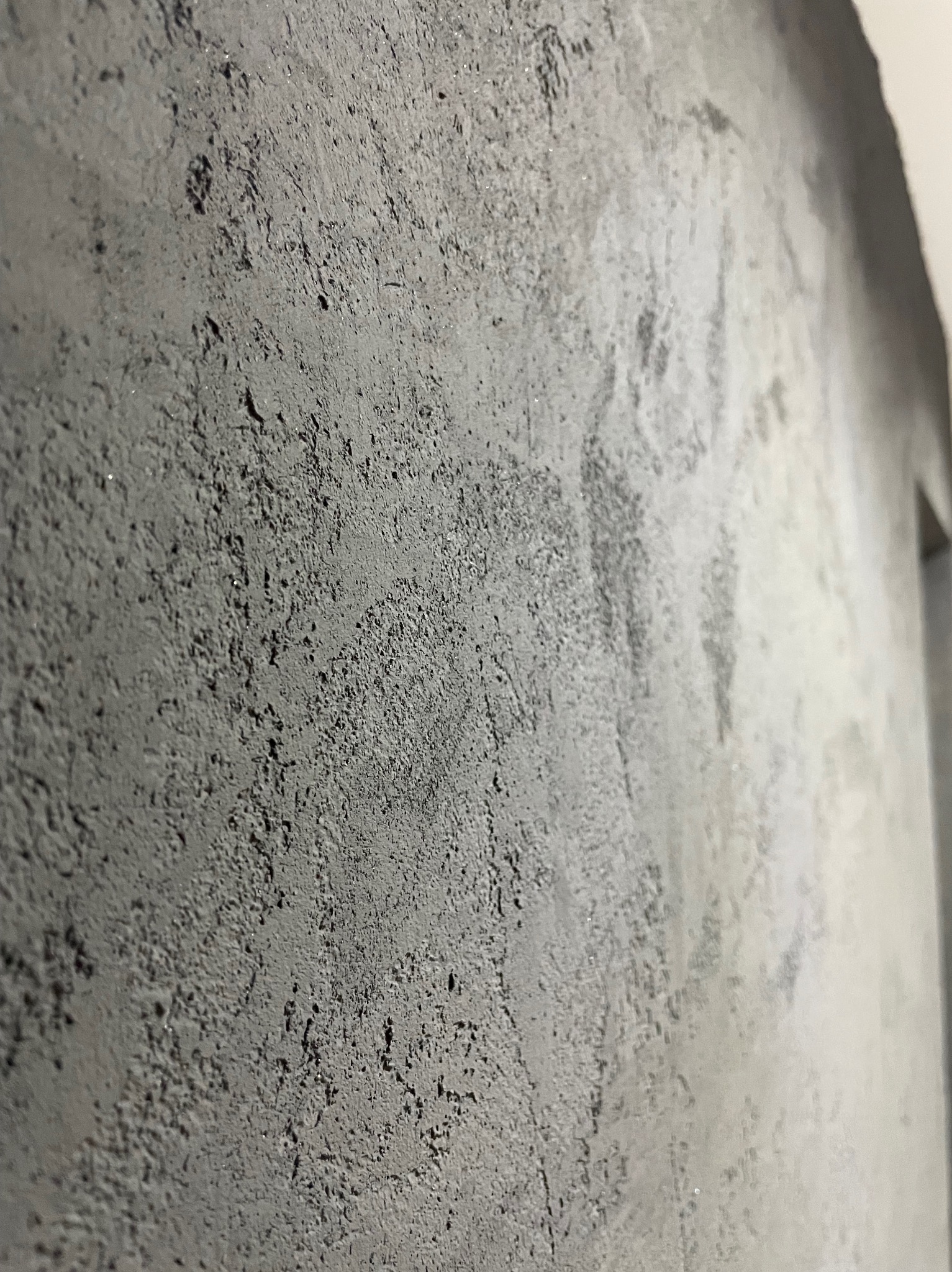 Szara ściana z wyraźną, chropowatą fakturą, uchwycona pod kątem, z widocznymi nieregularnościami i subtelnymi refleksami światła.