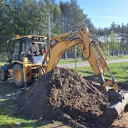Żółta koparko-ładowarka CAT z operatorem w kabinie, obok wykopu i sterty ziemi na tle ogrodzenia i drzew