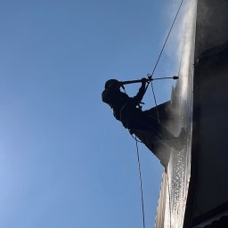 Krysztof Szewski ROOF Cleaning - Pracownik w uprzęży alpinistycznej czyści elewację budynku w Wadowicach za pomocą myjki ciśnieniowej, widok od dołu na tle błękitnego nieba.
