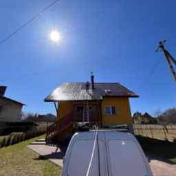 Pracownik na dachu żółtego domu, stojący na drabinie opierającej się o komin, widok zza białego samochodu dostawczego w słoneczny dzień.