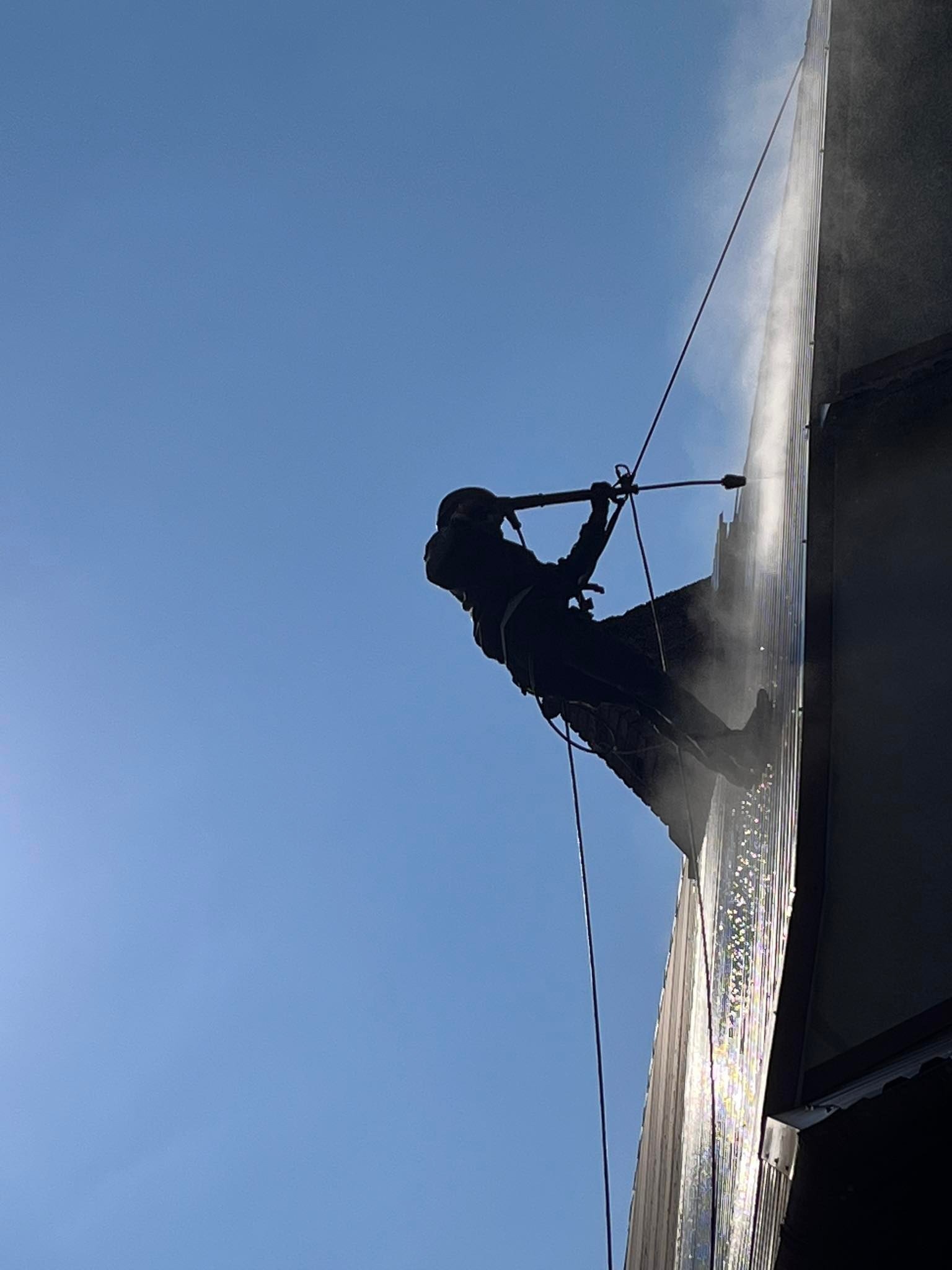 Pracownik w uprzęży alpinistycznej czyści elewację budynku w Wadowicach za pomocą myjki ciśnieniowej, widok od dołu na tle błękitnego nieba.