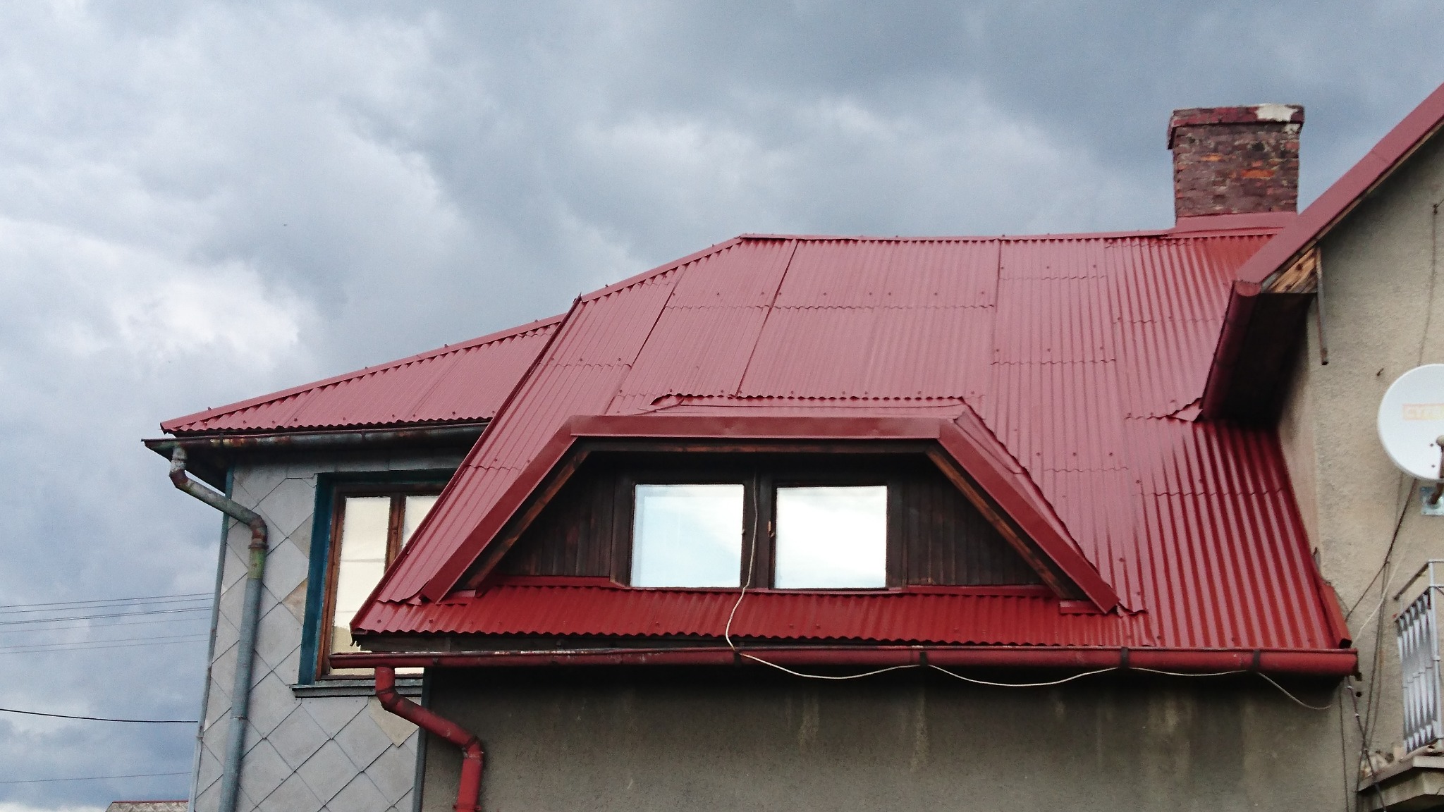Czerwony, blaszany dach z oknem dachowym i ceglanym kominem na tle pochmurnego nieba, widoczny fragment rynny i elewacji sąsiedniego budynku.