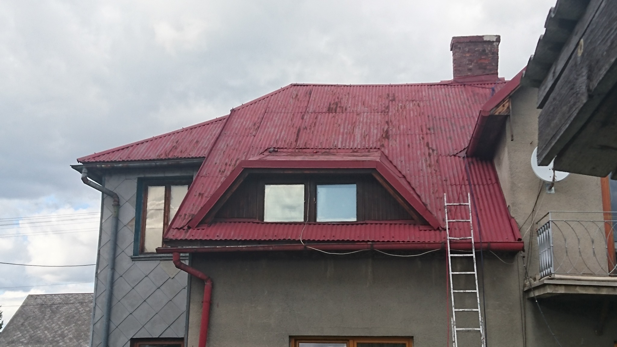 Czerwony, pofałdowany dach z lukarną i kominem, widziany z dołu, z drabiną opartą o ścianę, na tle pochmurnego nieba.