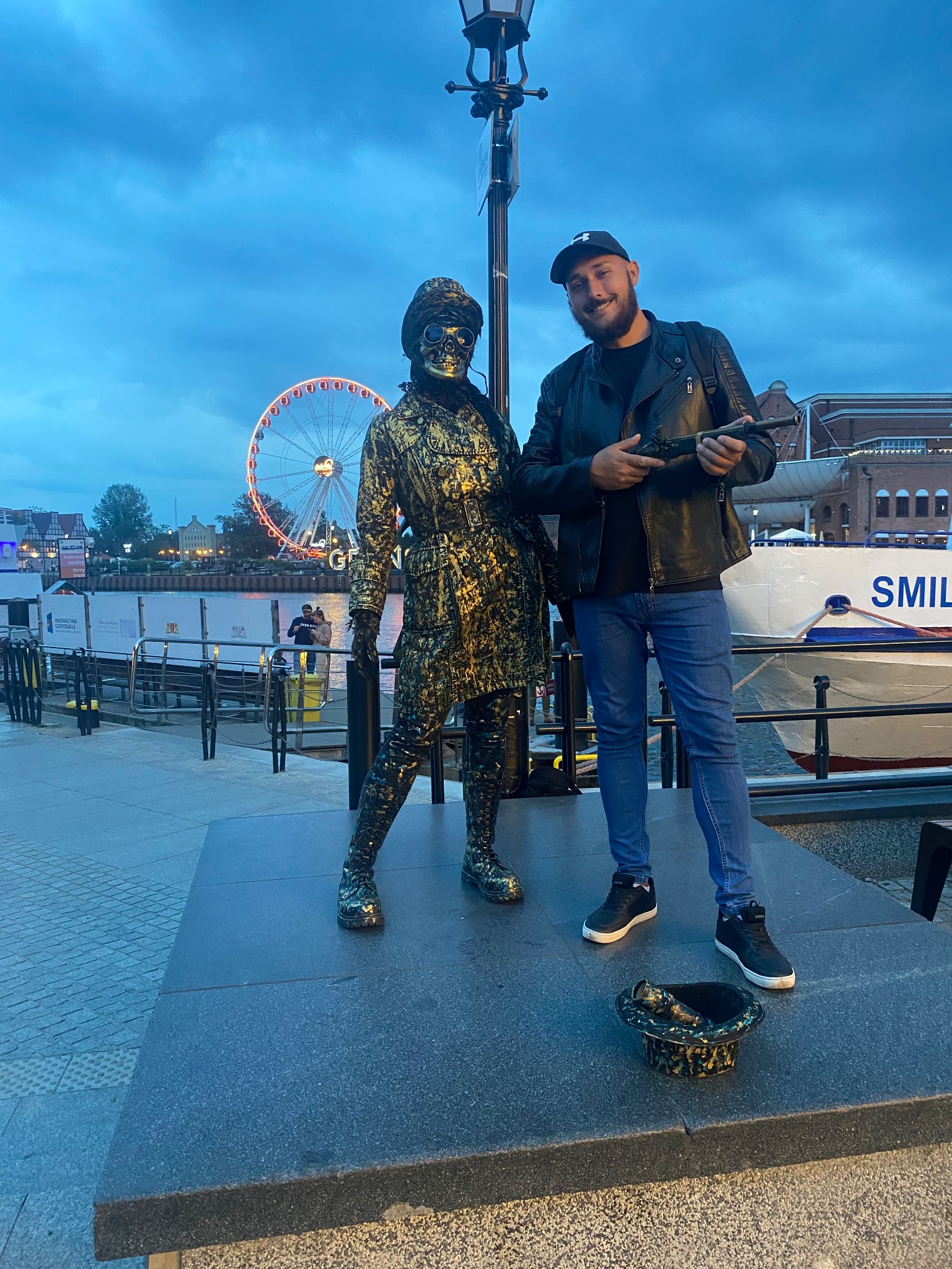Uśmiechnięty mężczyzna w skórzanej kurtce i czapce pozuje z flintą obok ulicznego performera w złotym kostiumie i masce, w tle diabelski młyn i budynki portowe w Gdańsku.
