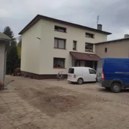 Dwukondygnacyjny dom po ociepleniu i pomalowaniu elewacji w kolorze kremowym z brązowym cokołem, zaparkowane samochody dostawcze i rower.