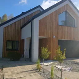 Nowoczesny dom jednorodzinny z elewacją z jasnego drewna, czarną stolarką okienną i bramą garażową, oraz białymi elementami wykończeniowymi, otoczony zielenią i kostką brukową.