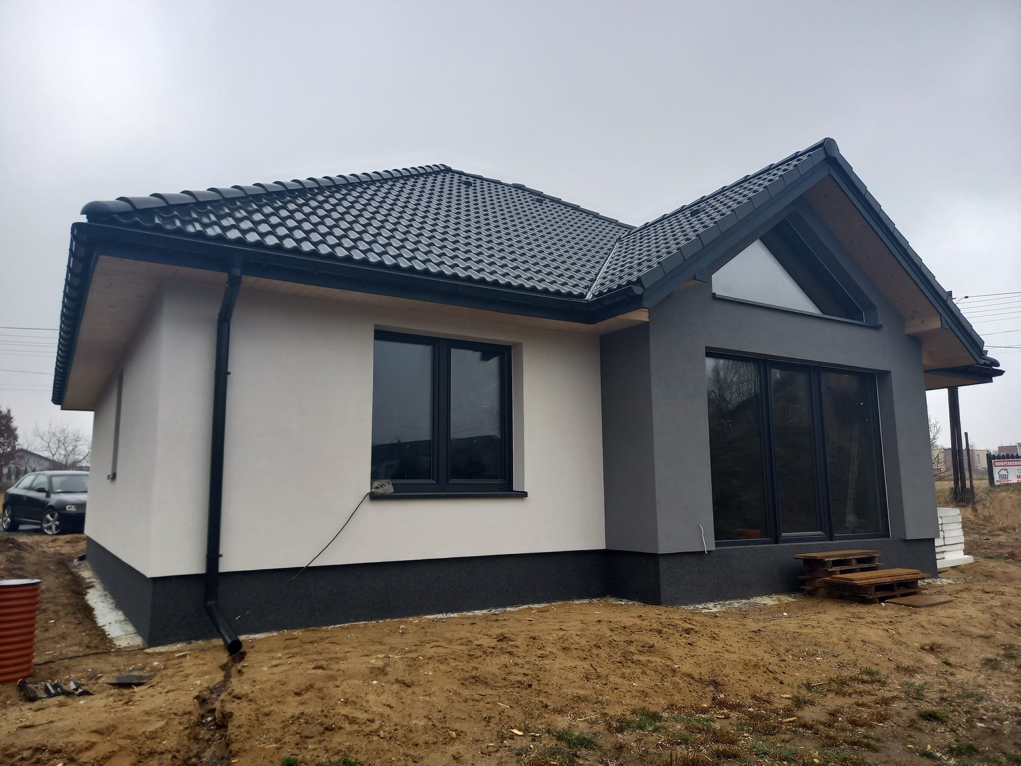 Nowoczesny dom jednorodzinny z dwukolorową elewacją: biały tynk i szara część z oknem trójkątnym, czarna dachówka i orynnowanie, widoczne palety jako prowizoryczne schody.