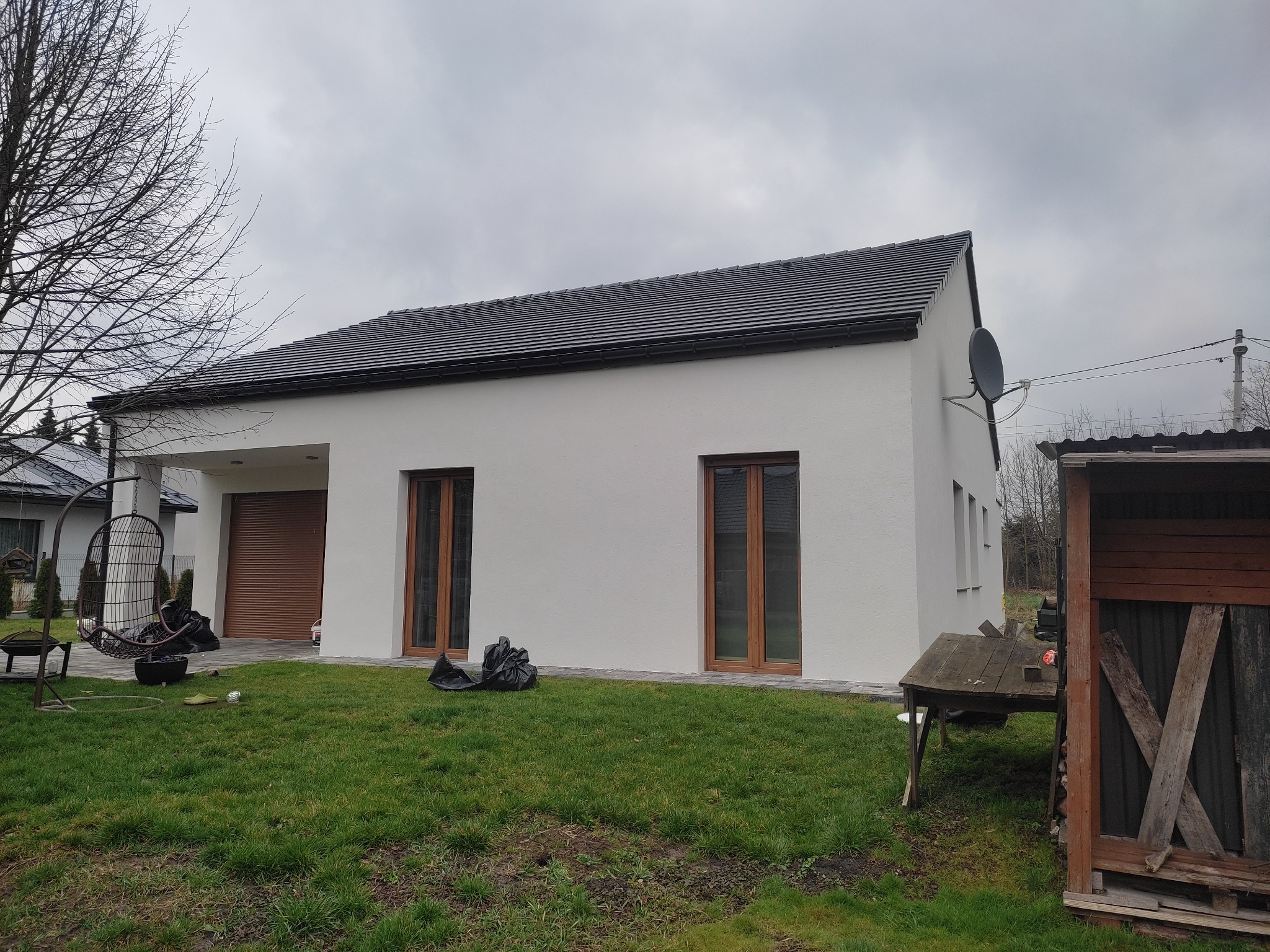 Nowoczesny dom z białą elewacją, ciemną dachówką i drewnianymi oknami, widoczne elementy zagospodarowania terenu: trawnik, huśtawka, altanka.