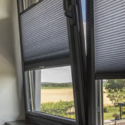 Szare, plisowane osłony okienne zamontowane w oknie z widokiem na pole i zieleń. Jedna z osłon częściowo rozsunięta, ukazująca krajobraz.