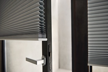 Szare, harmonijkowe osłony okienne typu duoplisy zamontowane w oknie z ciemną ramą i srebrną klamką, widoczne z bliska.