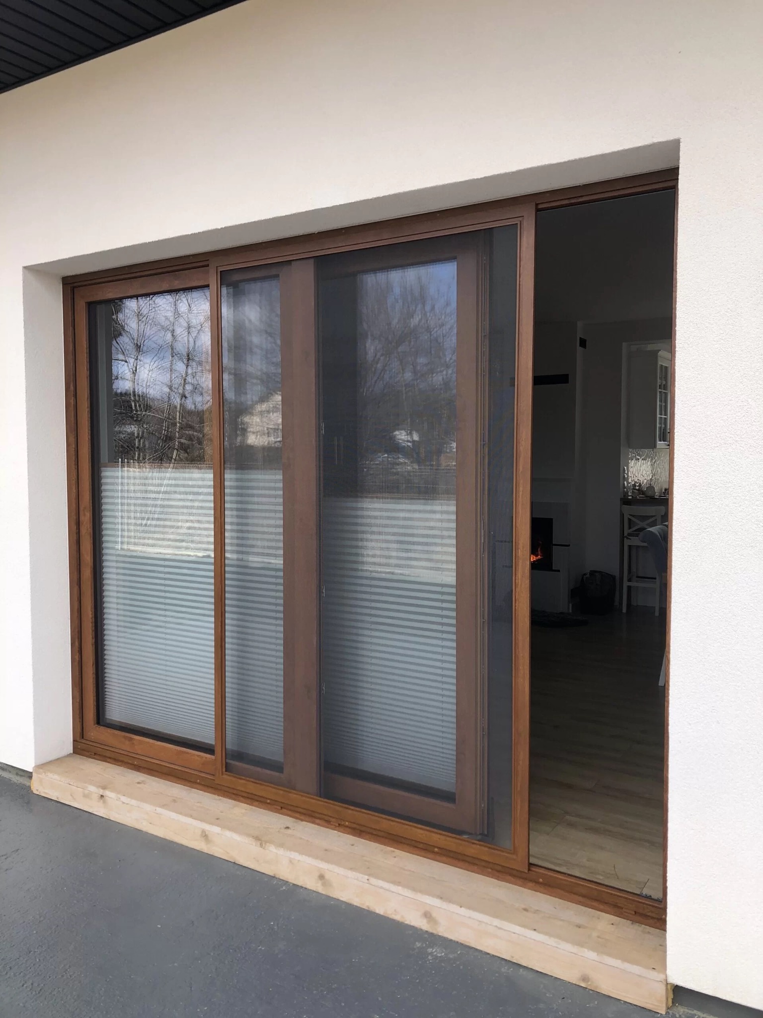 Przesuwne drzwi tarasowe z brązowymi ramami, wyposażone w plisowane rolety w oknach i moskitierę w otwartym skrzydle, widok na wnętrze domu z kominkiem.