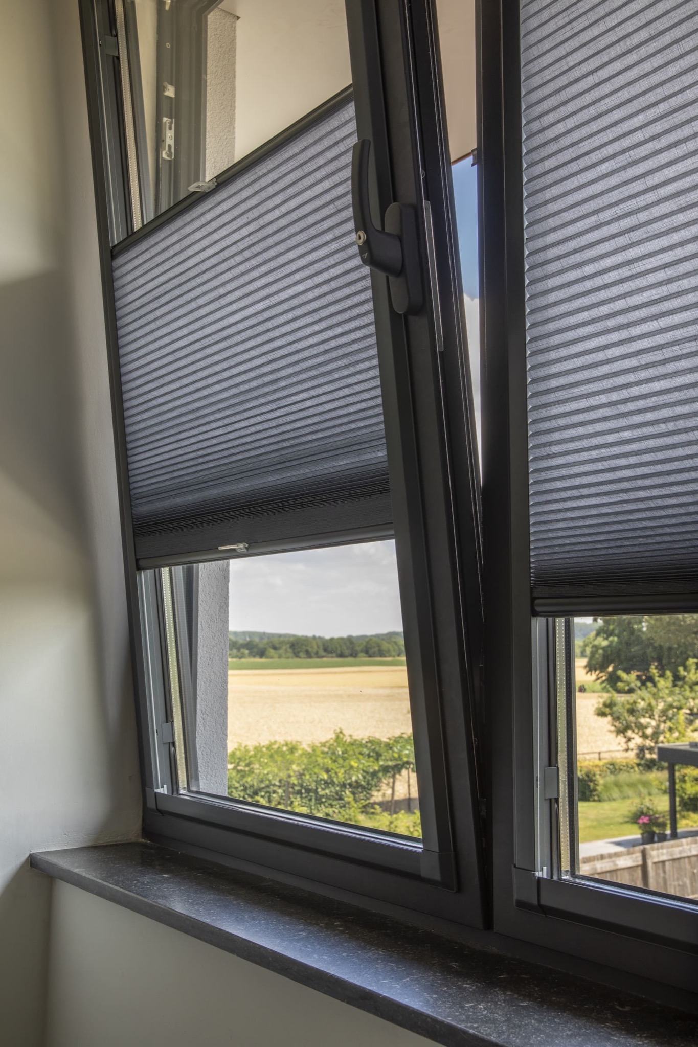 Szare, plisowane osłony okienne zamontowane w oknie z widokiem na pole i zieleń. Jedna z osłon częściowo rozsunięta, ukazująca krajobraz.