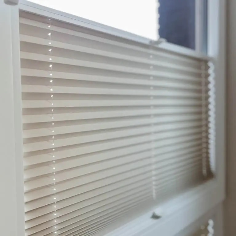 Beżowe, plisowane osłony okienne zamontowane w białej ramie okiennej, widoczne z bliskiej perspektywy, z delikatnym światłem wpadającym przez szczeliny.