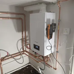 Nowoczesny biały piec gazowy marki Remeha z wyświetlaczem, podłączony miedzianymi rurami, z analizatorem spalin i czujnikiem temperatury zamontowanymi obok, w surowym pomieszczeniu.