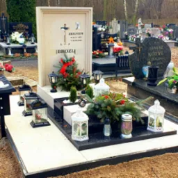 Jasny nagrobek z wyciętym krzyżem i zdjęciem osoby zmarłej, udekorowany zielonymi gałązkami, czerwonymi jagodami i lampionami, na tle innych grobów na cmentarzu.