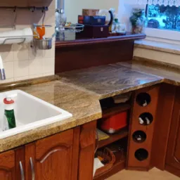 Fragment kuchni z brązowymi szafkami i blatem w odcieniach beżu i brązu, zlewozmywak z baterią, wiszące przybory kuchenne, okno z widokiem na zimowy krajobraz.