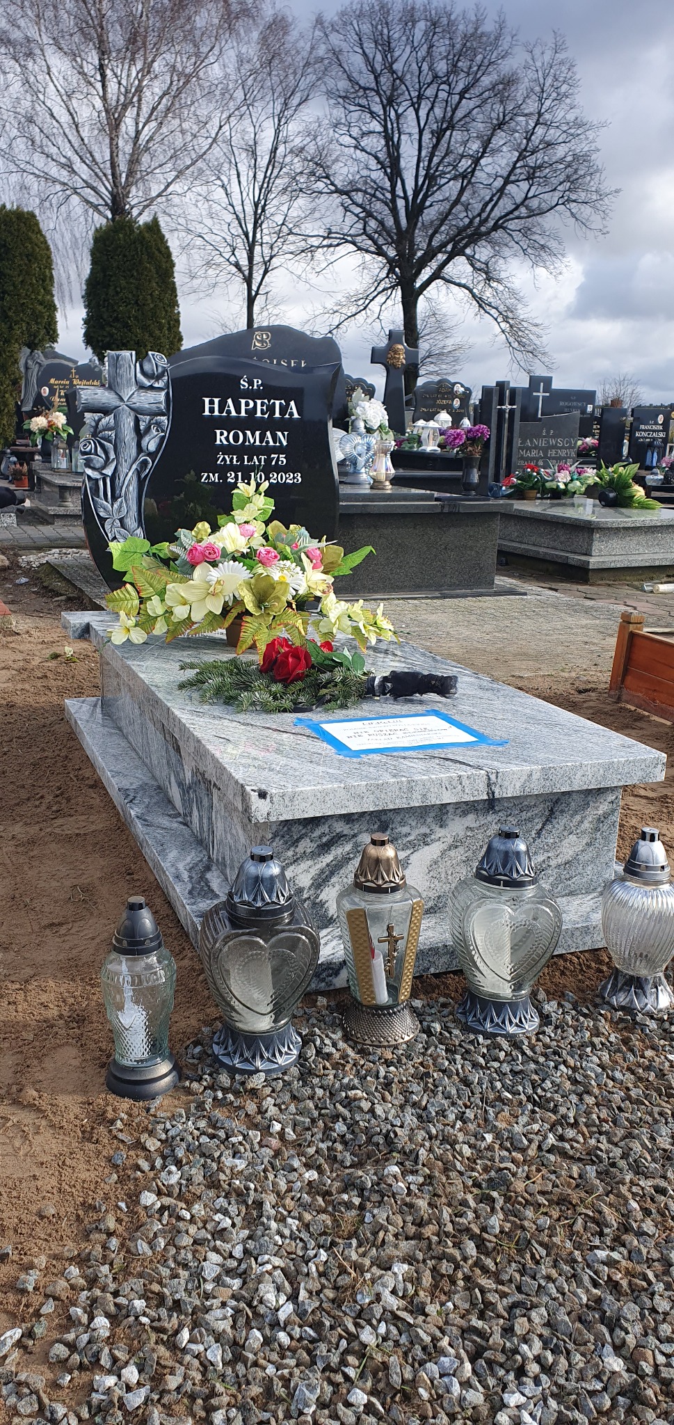 Granitowy nagrobek z czarną tablicą i kwiatami, otoczony lampionami na żwirowym podłożu, w tle inne groby i drzewa na cmentarzu.