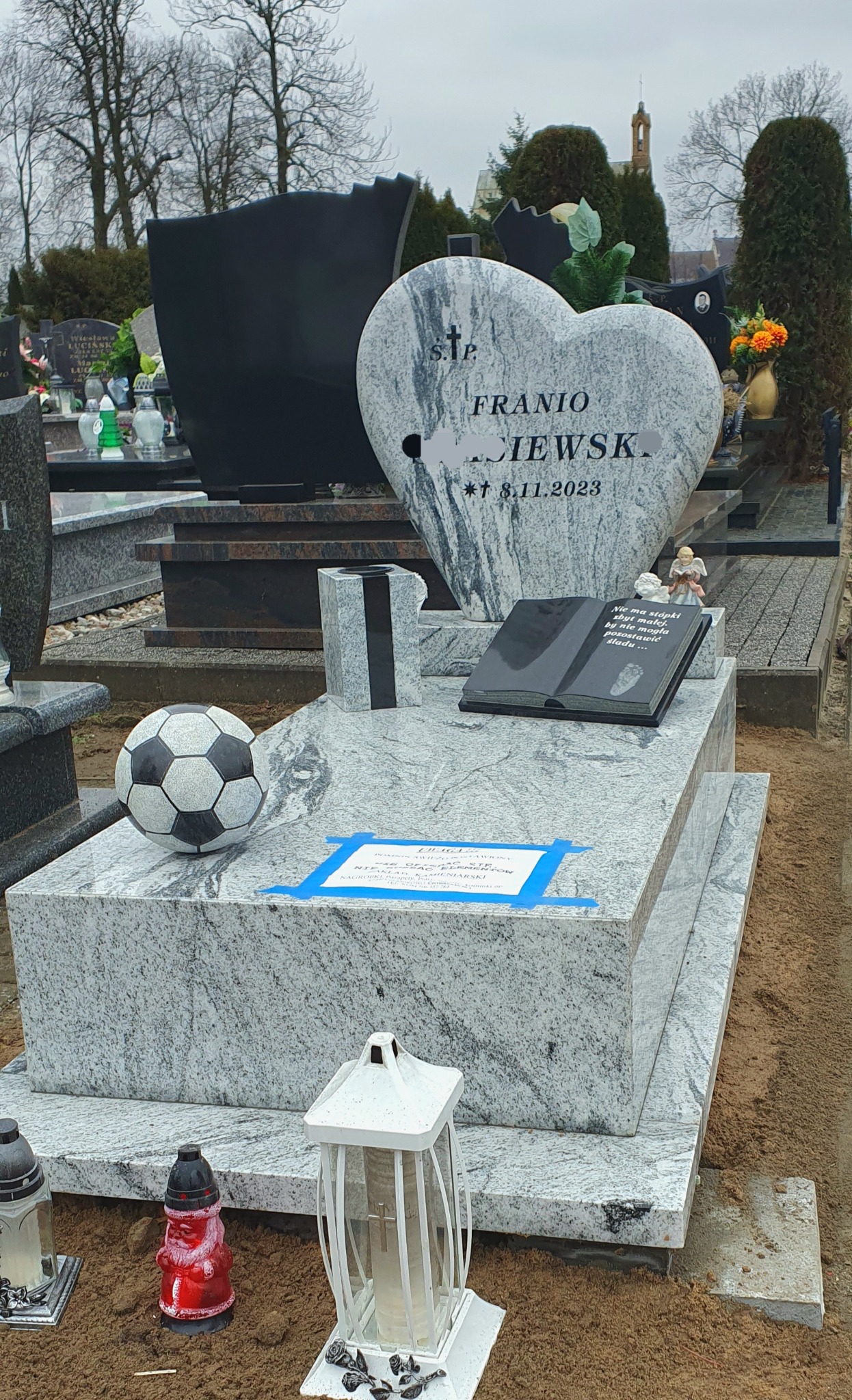 Jasnoszary, granitowy nagrobek z sercem i książką, ozdobiony piłką nożną i lampionami na cmentarzu. Na nagrobku wyryte imię i nazwisko oraz data 8.11.2023. Widoczny fragment kartki z napisem...