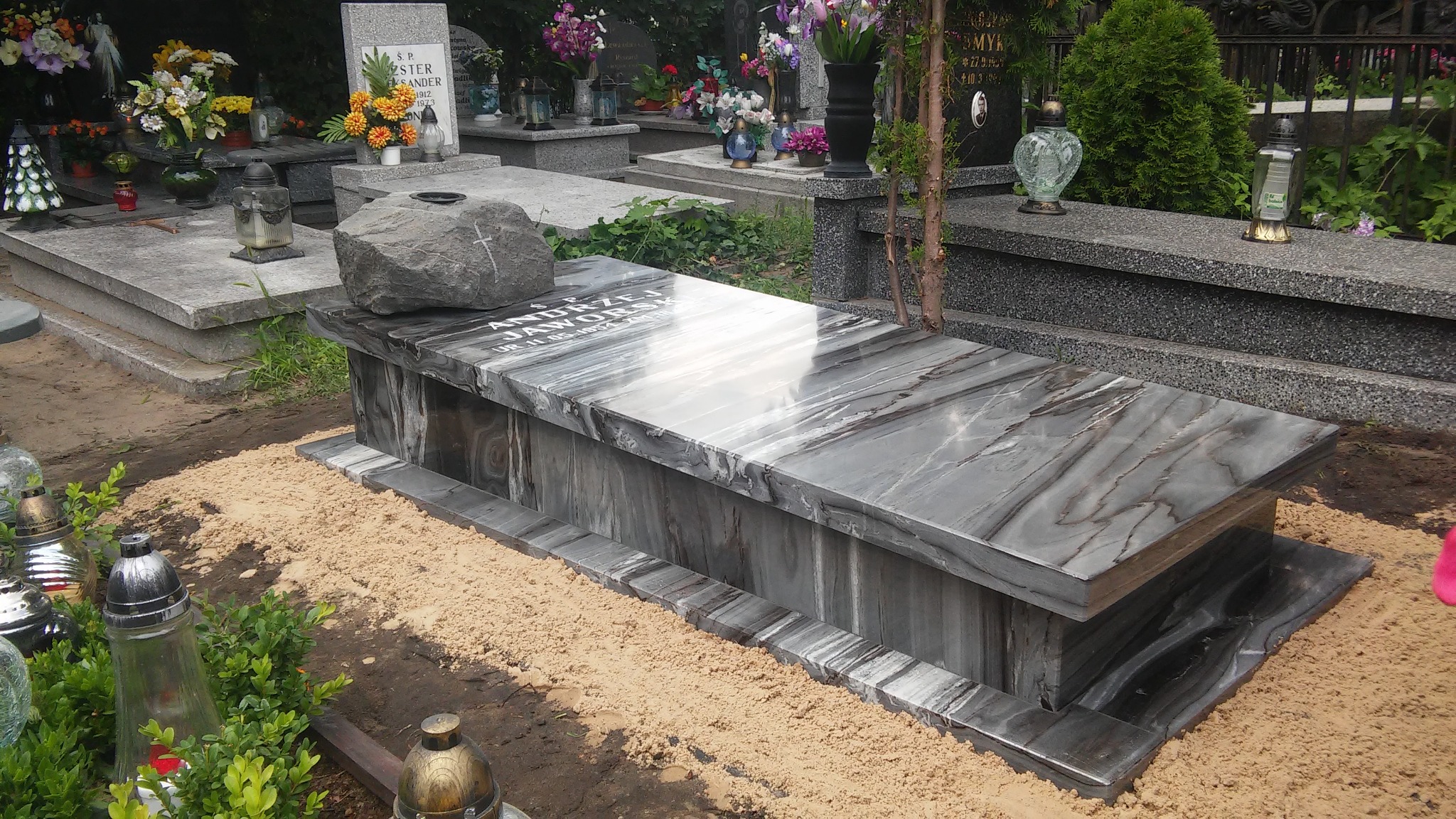 Nowoczesny nagrobek z szarego kamienia z wyraźnym wzorem żyłkowania, przykryty dużym, surowym kamieniem z wyrytym krzyżem, otoczony piaskiem i innymi nagrobkami na cmentarzu.