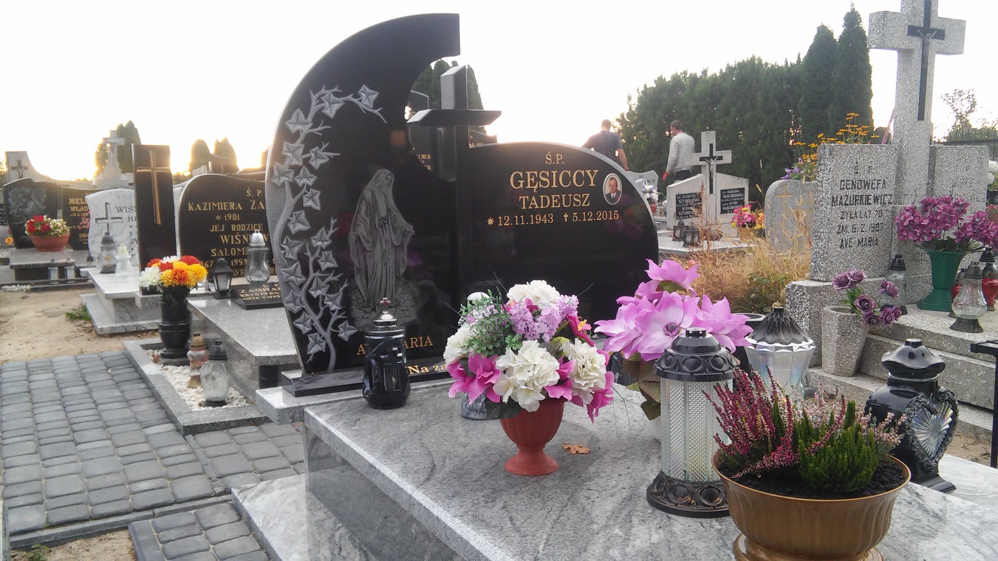 Czarny nagrobek z wygrawerowaną postacią i datami, ozdobiony kwiatami w wazonie na granitowej płycie, na tle cmentarza z innymi nagrobkami i zielenią.