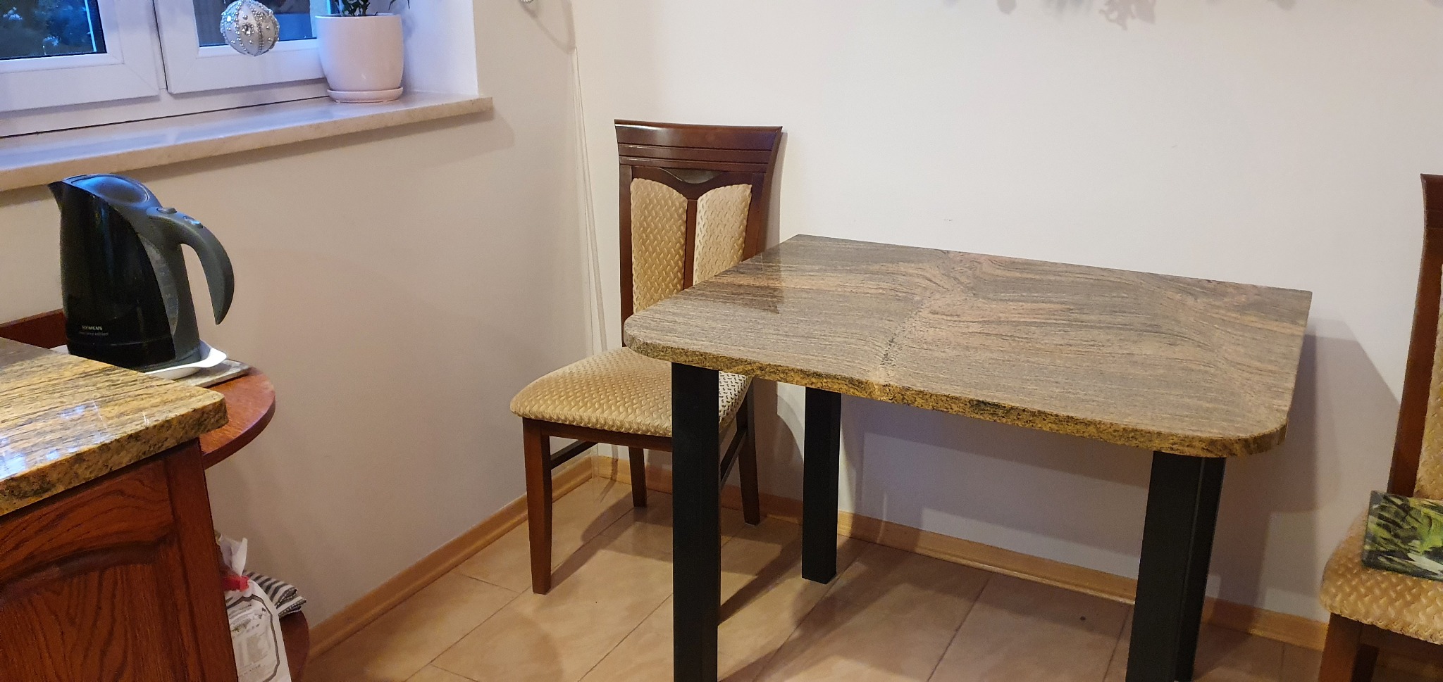 Stół z blatem z kamienia o nieregularnym kształcie, wsparty na czarnych nogach, w tle krzesła z tapicerką w odcieniach beżu.