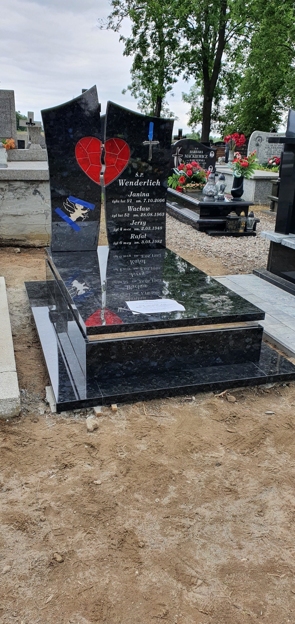 Czarny, polerowany nagrobek z motywem pękniętego czerwonego serca i wygrawerowanym wizerunkiem anioła, umieszczony na cmentarzu.