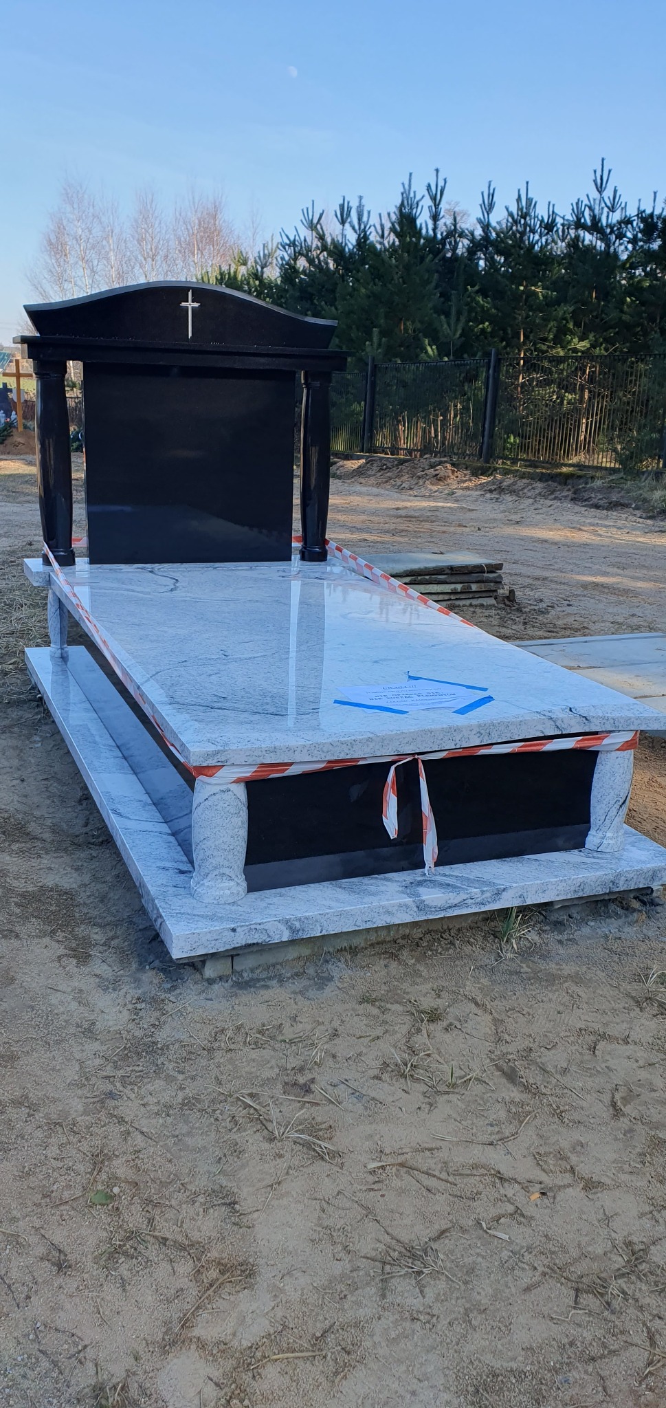 Nowy, jasnoszary nagrobek z czarnym panelem i kolumnami, zabezpieczony taśmą ostrzegawczą, na tle drzew i nieba.