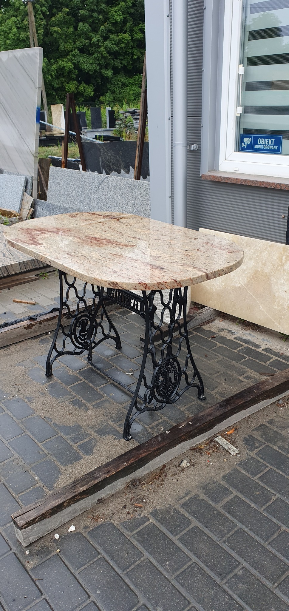 Owalny blat stołu z jasnego kamienia z czerwonymi żyłkami, osadzony na czarnej, ozdobnej podstawie od maszyny do szycia Singer, na tle magazynu kamieniarskiego.