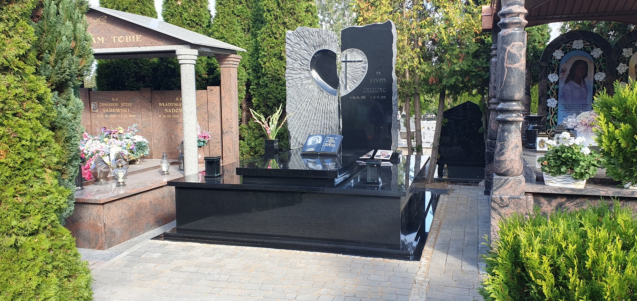 Czarny, nowoczesny pomnik z elementami rzeźbionymi w kształcie serca i krzyża, otoczony zielenią cmentarza i innymi nagrobkami.