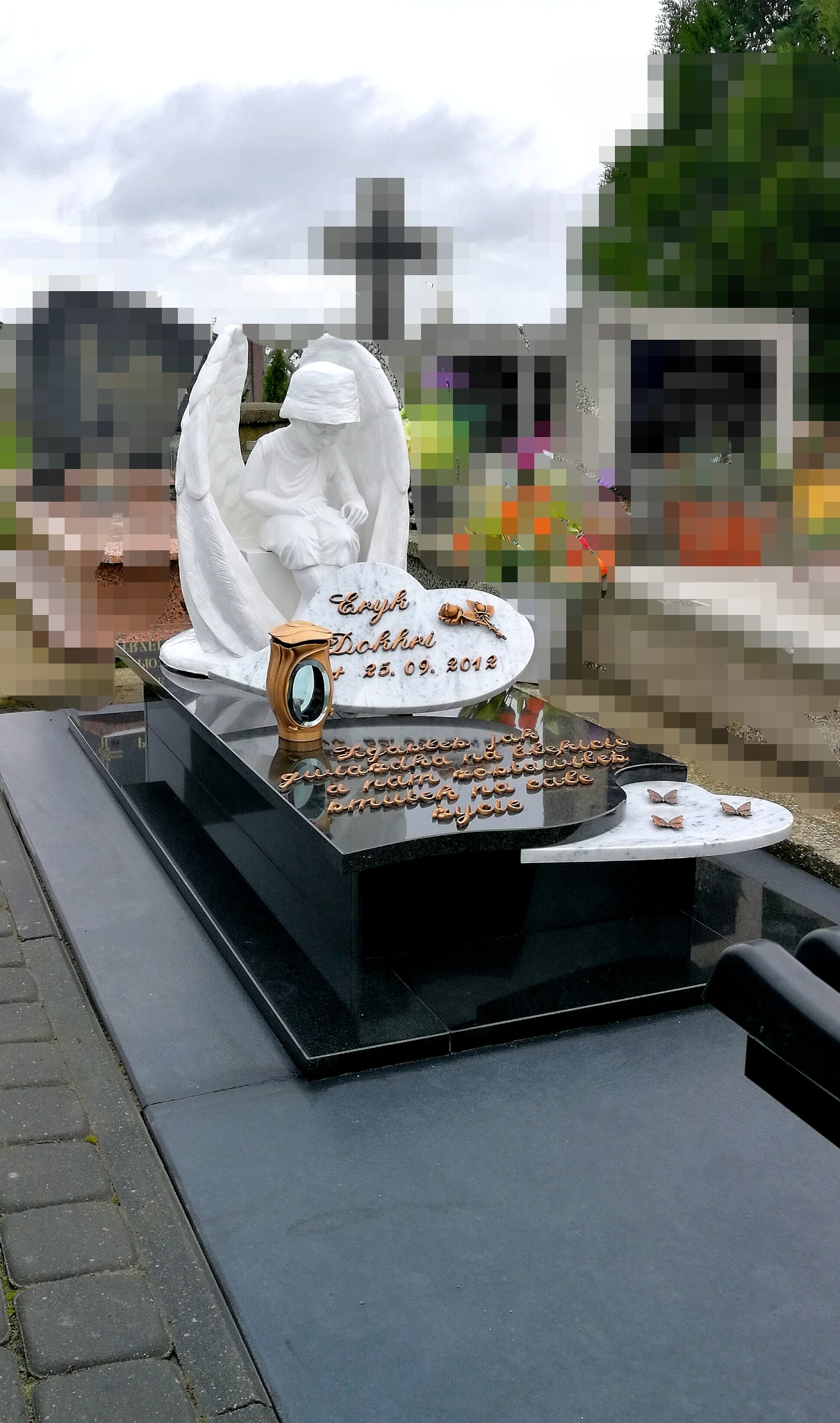 Czarny, błyszczący pomnik z białym marmurowym dodatkiem w kształcie chmury z wygrawerowanym imieniem i datą, zwieńczony rzeźbą anioła, w tle krzyż i inne nagrobki.