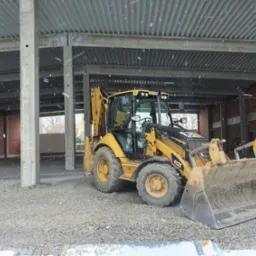 Żółta koparko-ładowarka Caterpillar 432E na placu budowy z widocznymi betonowymi kolumnami i stalową konstrukcją dachu, lekko ośnieżonym podłożem z gruzu i padającym śniegiem.