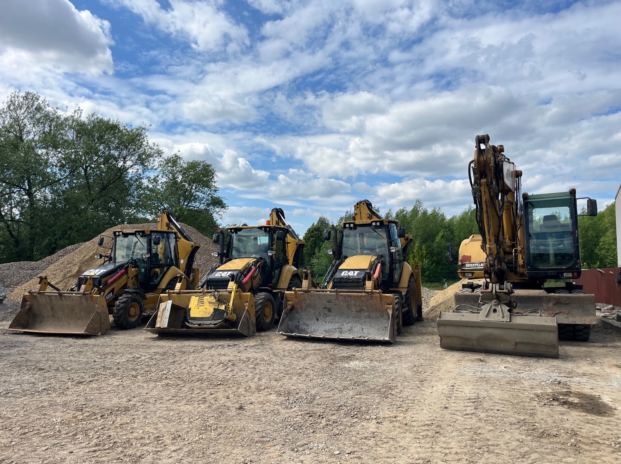 Żółte koparko-ładowarki CAT ustawione rzędem na żwirowym placu, z hałdą ziemi i zielenią w tle, pod błękitnym niebem z obłokami.