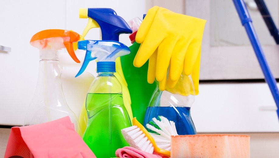 Zestaw środków czystości: butelki z rozpylaczami, płyny w różnych kolorach, szczotka z białym włosiem, gąbka i gumowe rękawice w odcieniach żółci i różu, na tle białych szafek.