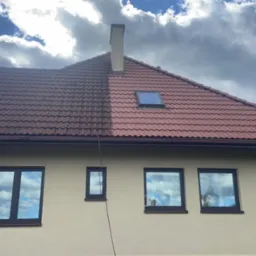 Częściowo wyczyszczony dach z czerwonej dachówki, widoczna różnica w kolorze po lewej i prawej stronie, beżowa elewacja domu, okna z odbiciem chmur, drabina oparta o ścianę.