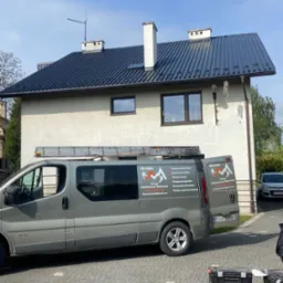 Dom z ciemnym, odnowionym dachem, drabina na dachu szarego vana firmy remontowej z logo i numerem telefonu, skrzynki narzędziowe na brukowanym podjeździe.