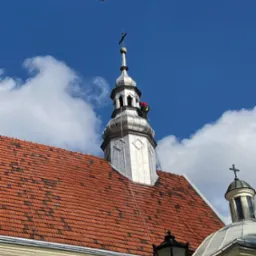 Praca wysokościowa na wieży kościoła z czerwoną dachówką. Pracownik w czerwonym kombinezonie zabezpieczony linami. Błękitne niebo z chmurami w tle.