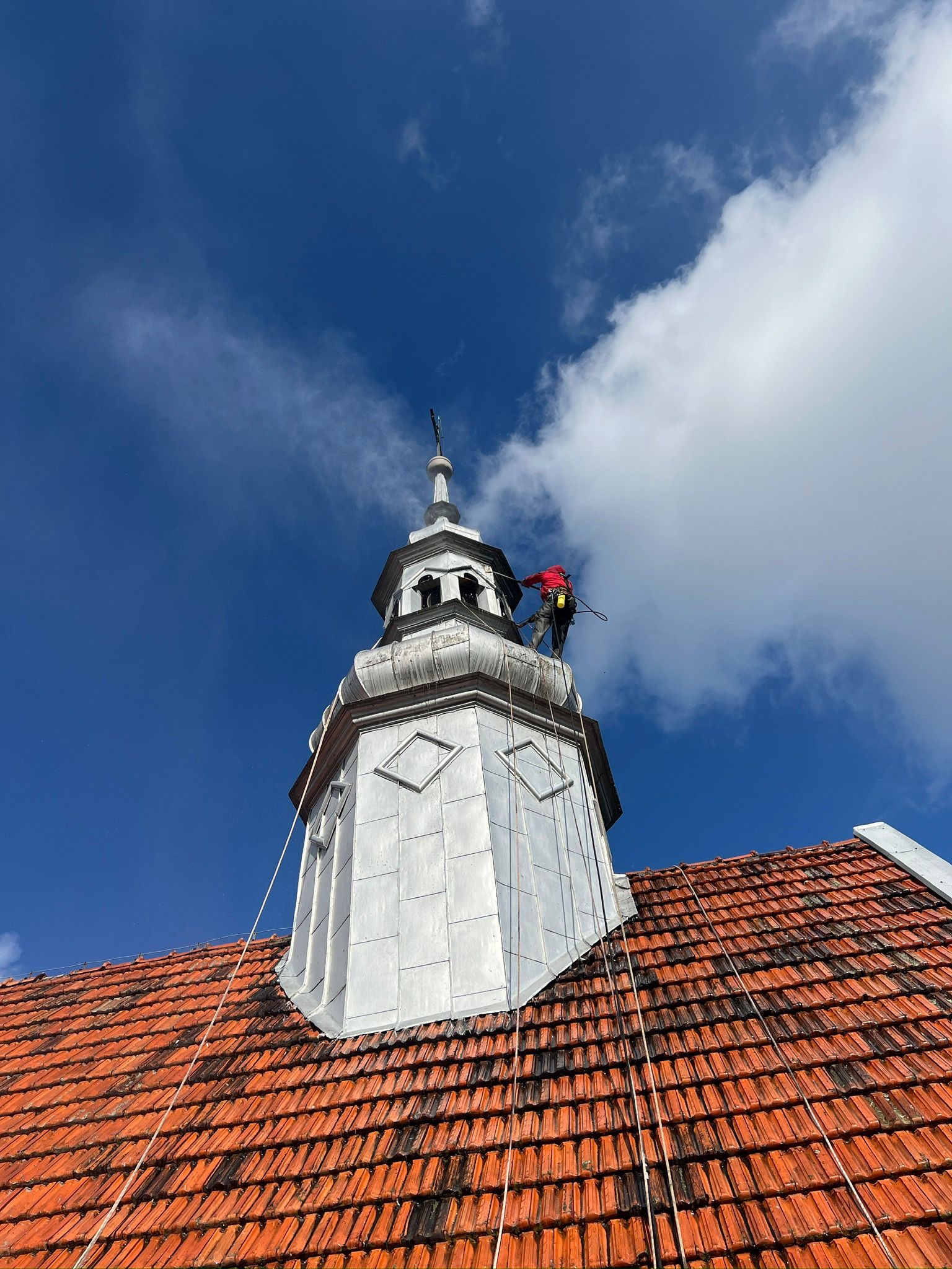 Pracownik w czerwonej kurtce, zabezpieczony linami, wykonuje prace na szczycie wieży kościoła z pomalowaną na szaro elewacją, na tle błękitnego nieba z białymi chmurami.