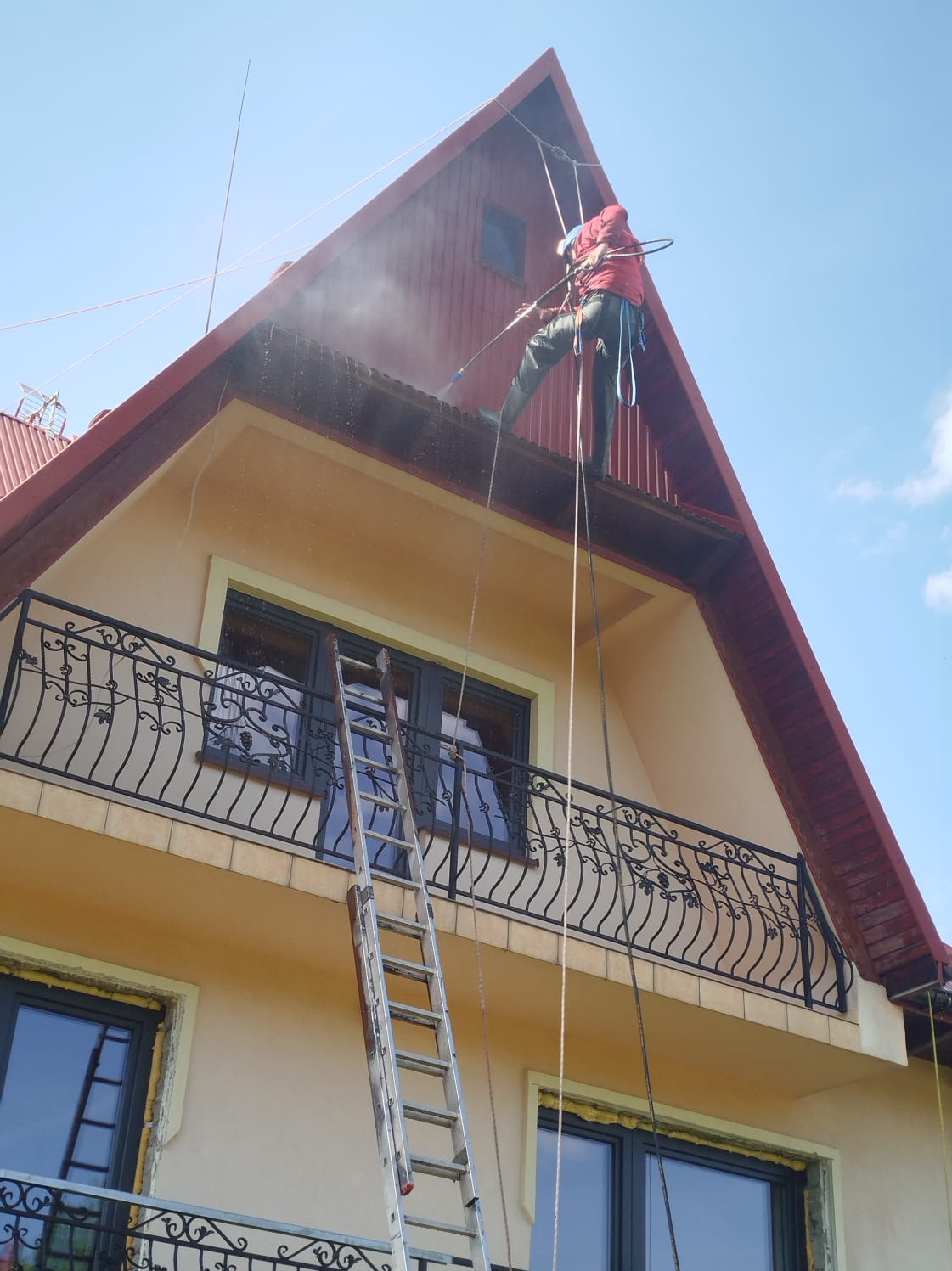 Pracownik w czerwonej kurtce myje dach domu z drabiny linowej, używając myjki ciśnieniowej. W tle widoczne jasne niebo i fragment balkonu z ozdobną balustradą.