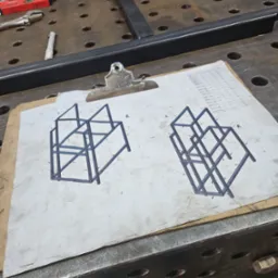 Szkic konstrukcji metalowej na desce z klipsem, obok częściowo zmontowana rama i narzędzia na stole spawalniczym.