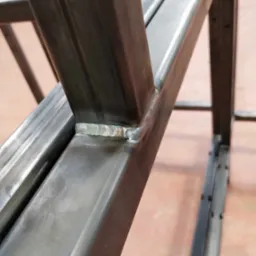 Szczegółowe ujęcie spawu łączącego metalowe profile konstrukcyjne pod kątem, widoczne ślady obróbki i niedoskonałości powierzchni.