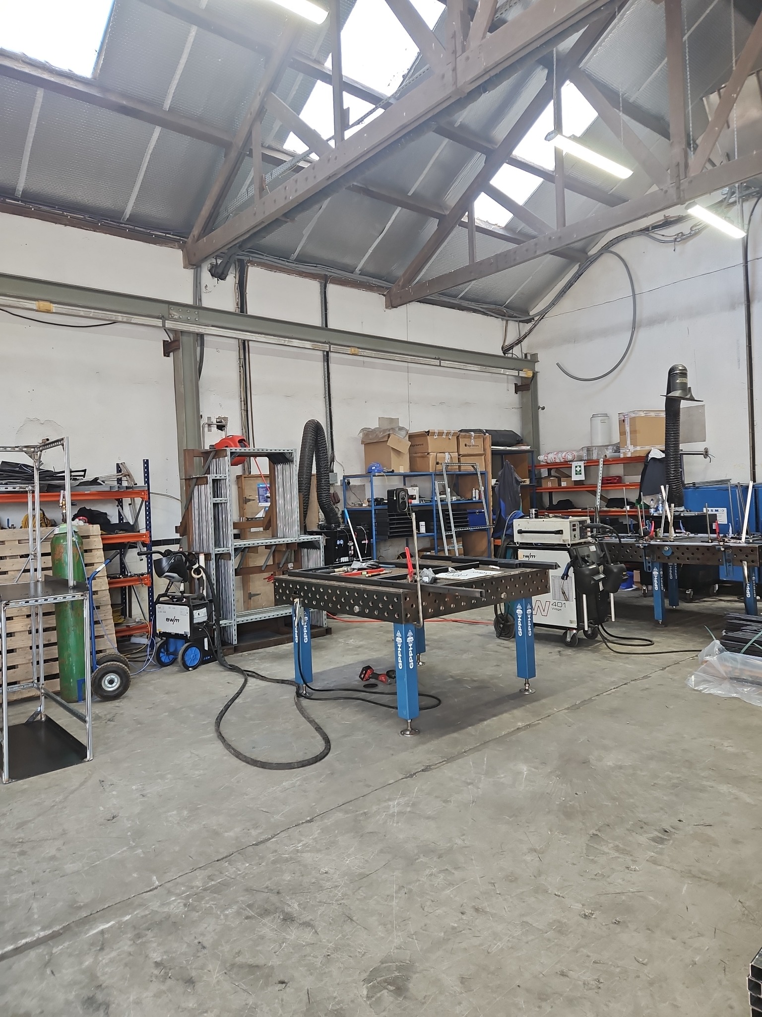 Wnętrze warsztatu z narzędziami i stołem spawalniczym, widoczne spawarki i materiały metalowe, oświetlenie górne, hala z konstrukcją stalową dachu.