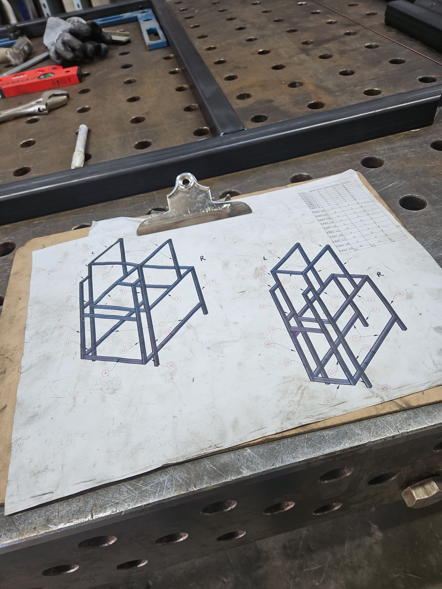 Szkic konstrukcji metalowej na desce z klipsem, obok częściowo zmontowana rama i narzędzia na stole spawalniczym.