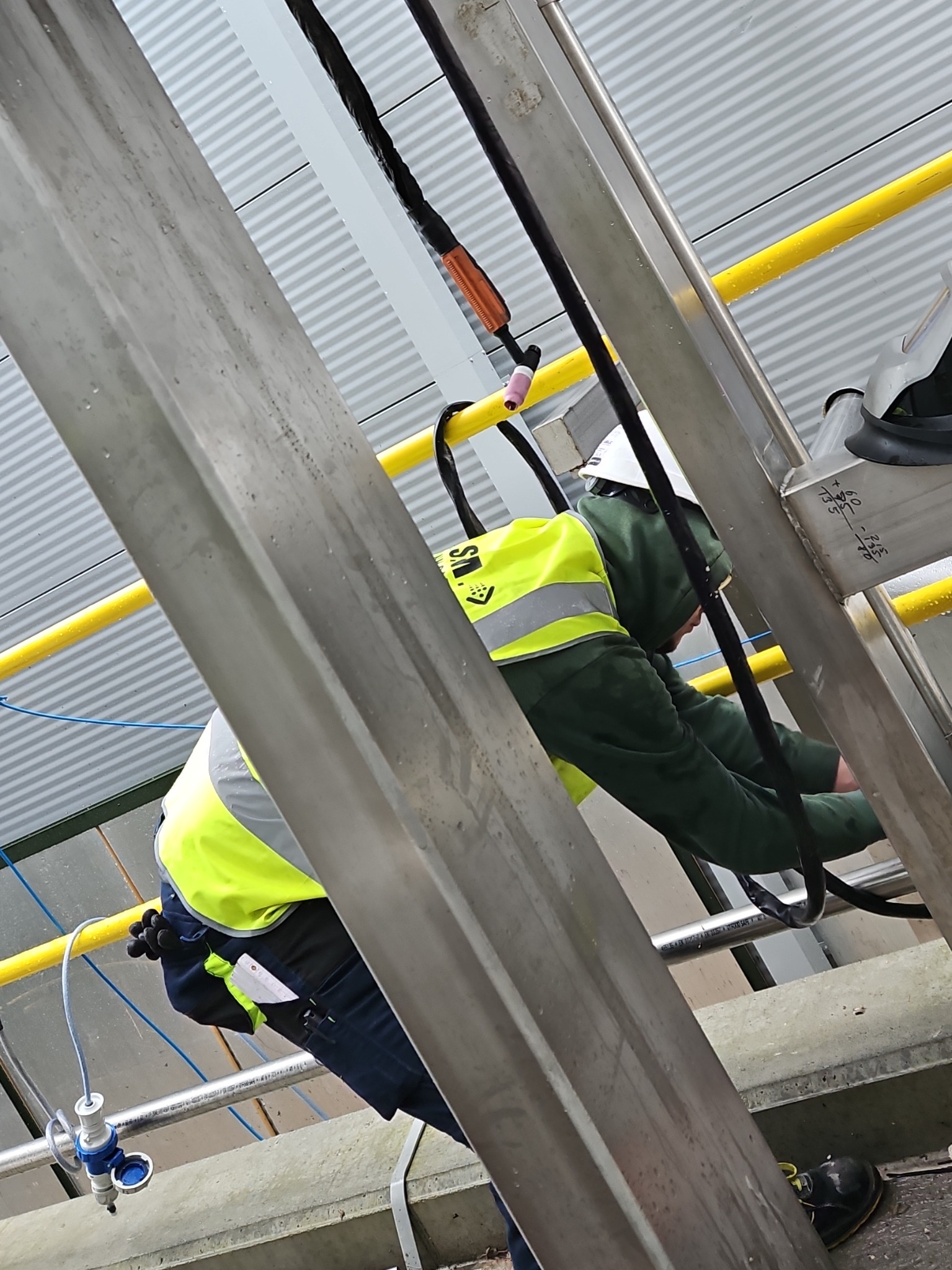 Pracownik w kasku i kamizelce odblaskowej, pochylony nad konstrukcją ze stali nierdzewnej, prawdopodobnie podczas prac spawalniczych lub montażowych na hali. Widoczne przewody i elementy spawarki.