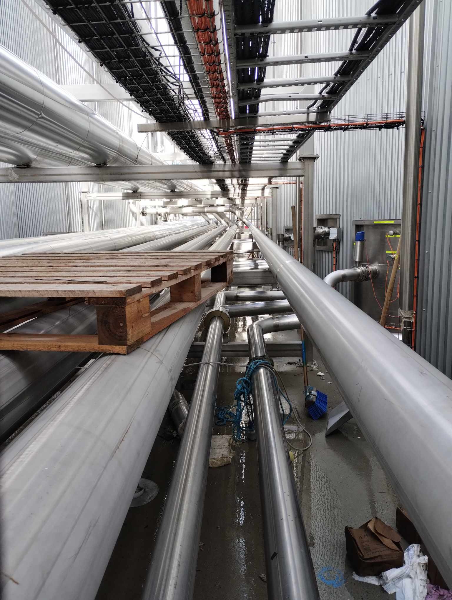 Wnętrze hali produkcyjnej z rzędami metalowych rur i koryt kablowych pod sufitem, drewniana paleta położona na rurach, widoczne ślady po budowie i montażu.